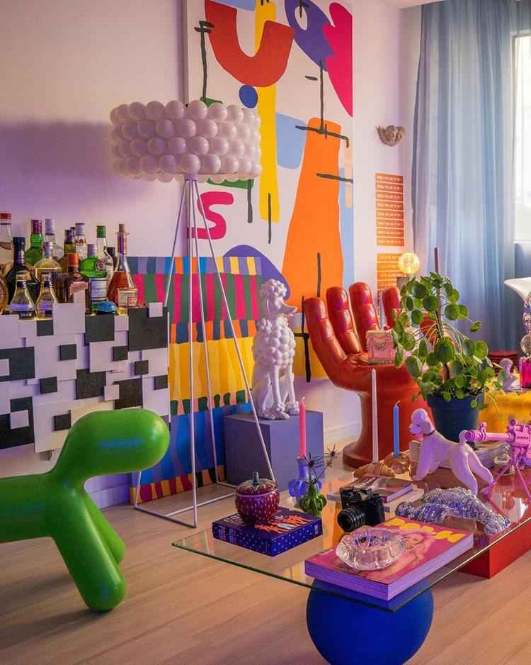 sala colorida com decoração maximalista, canto com paredes brancas, chão de madeira, mesa de vidro ao centro com diversos objetos