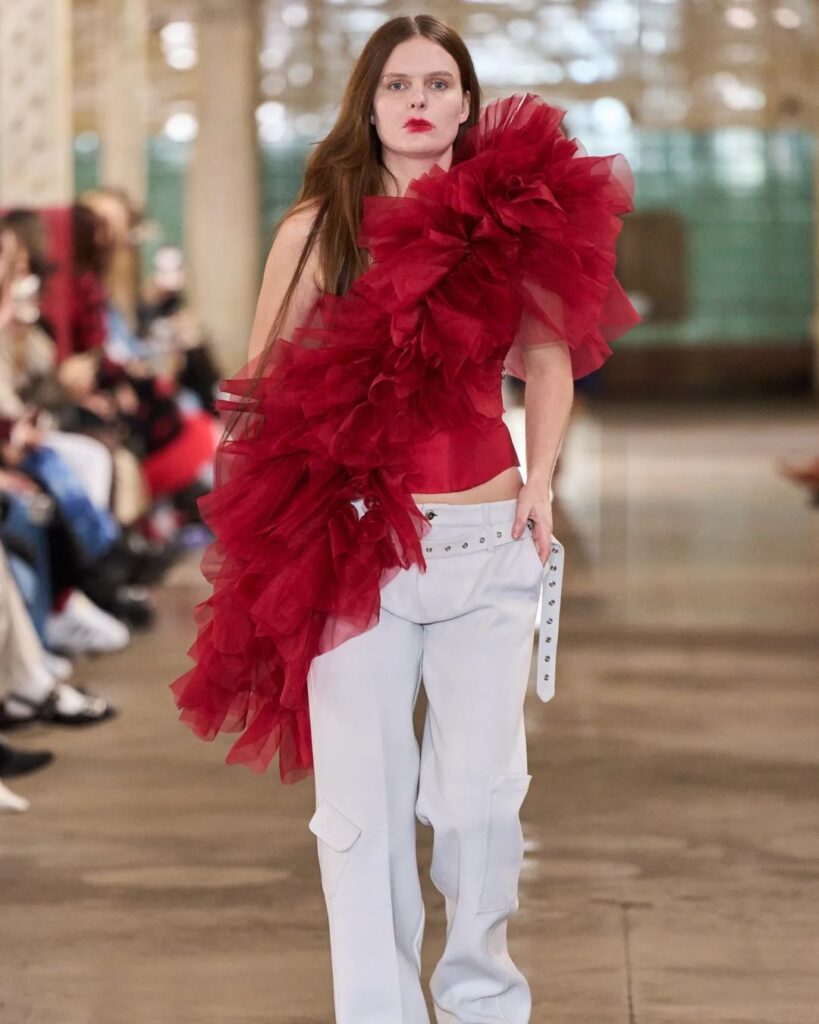 Foto do desfile da Marques’Almeida na London Fashion Week. Na imagem, há uma mulher branca vestindo uma blusa com babados vermelhos e uma calça cargo branca. 
