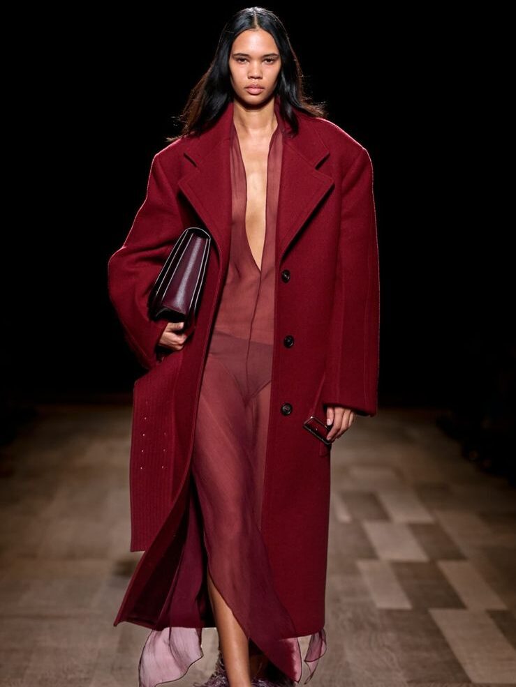 Ferragamo traz a tendência, com modelo que usa vestido vermelho bordô com transparência, sobreposto por um casaco de mesma cor e carrega uma bolsa, na mesma paleta, abaixo do braço.