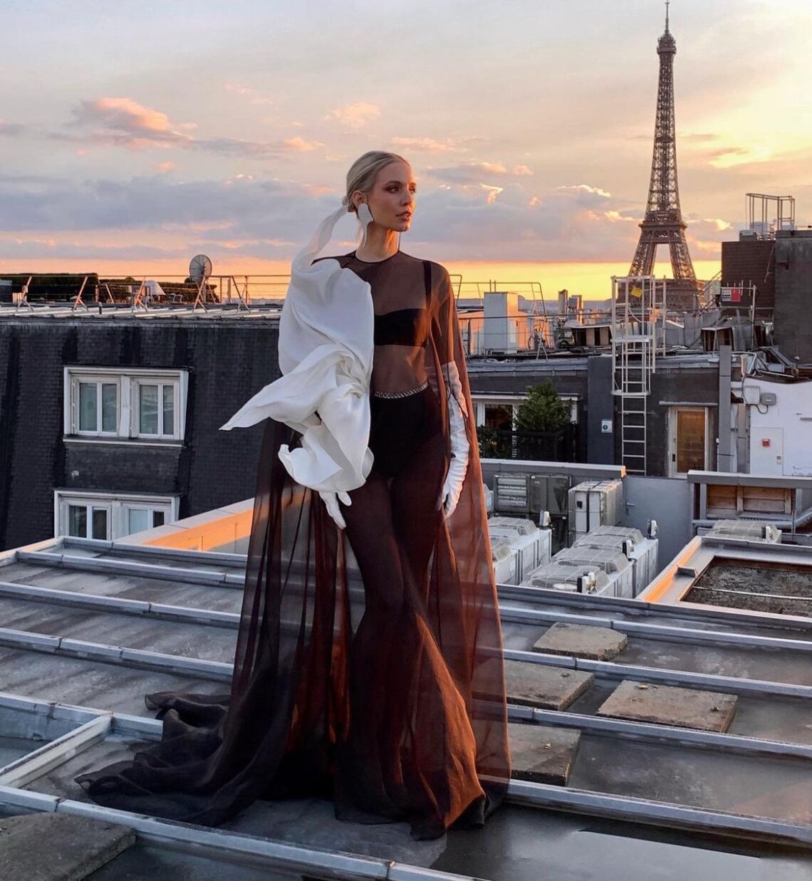 Leonie Hanne usa vestido transparente na cor marrom, com detalhe branco no ombro. Ela está em um telhado e atrás é possível ver a Torre Eiffel.