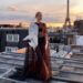 Leonie Hanne usa vestido transparente na cor marrom, com detalhe branco no ombro. Ela está em um telhado e atrás é possível ver a Torre Eiffel.