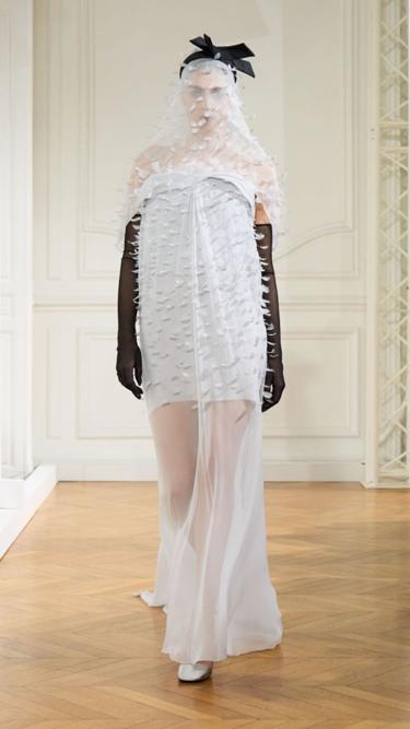 Modelo usa vestido branco com espécie de véu e bordados, desde a face até a altura das coxas. Complementa o visual com luvas na cor preta, e acessório com laço preto, no alto da cabeça, em desfile da Givenchy.