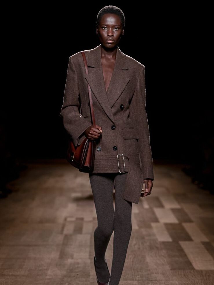 Modelo negra defila para a marca italiana Ferragamo, usando blazer e meia-calça cinzas, além de bolsa marrom abaixo do braço.