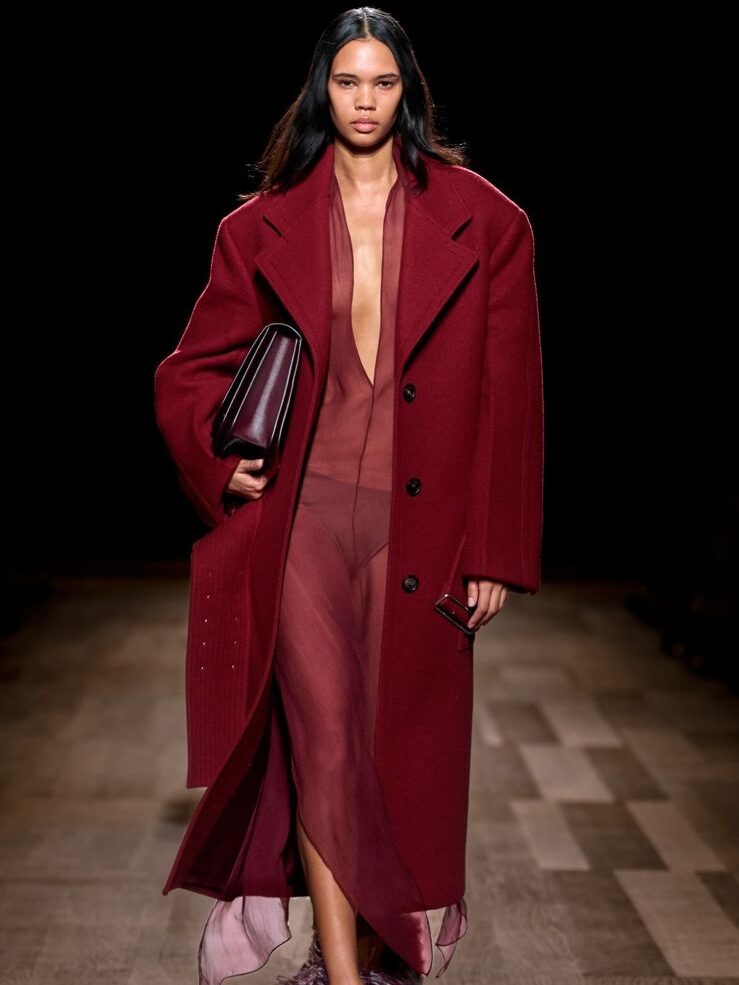 Modelo na passarela, desfilando com um casaco longo de lã pesada, por cima de um vestido transparente e levando abaixo dos braços uma bolsa-maleta. Todos os itens são na cor vermelha.