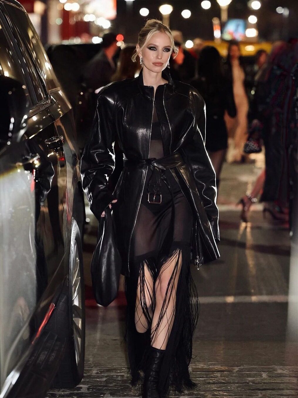 Leonie Hanne usando um vestido transparente com franjas, coturno e um casaco puffer de couro por cima, em um look todo preto.