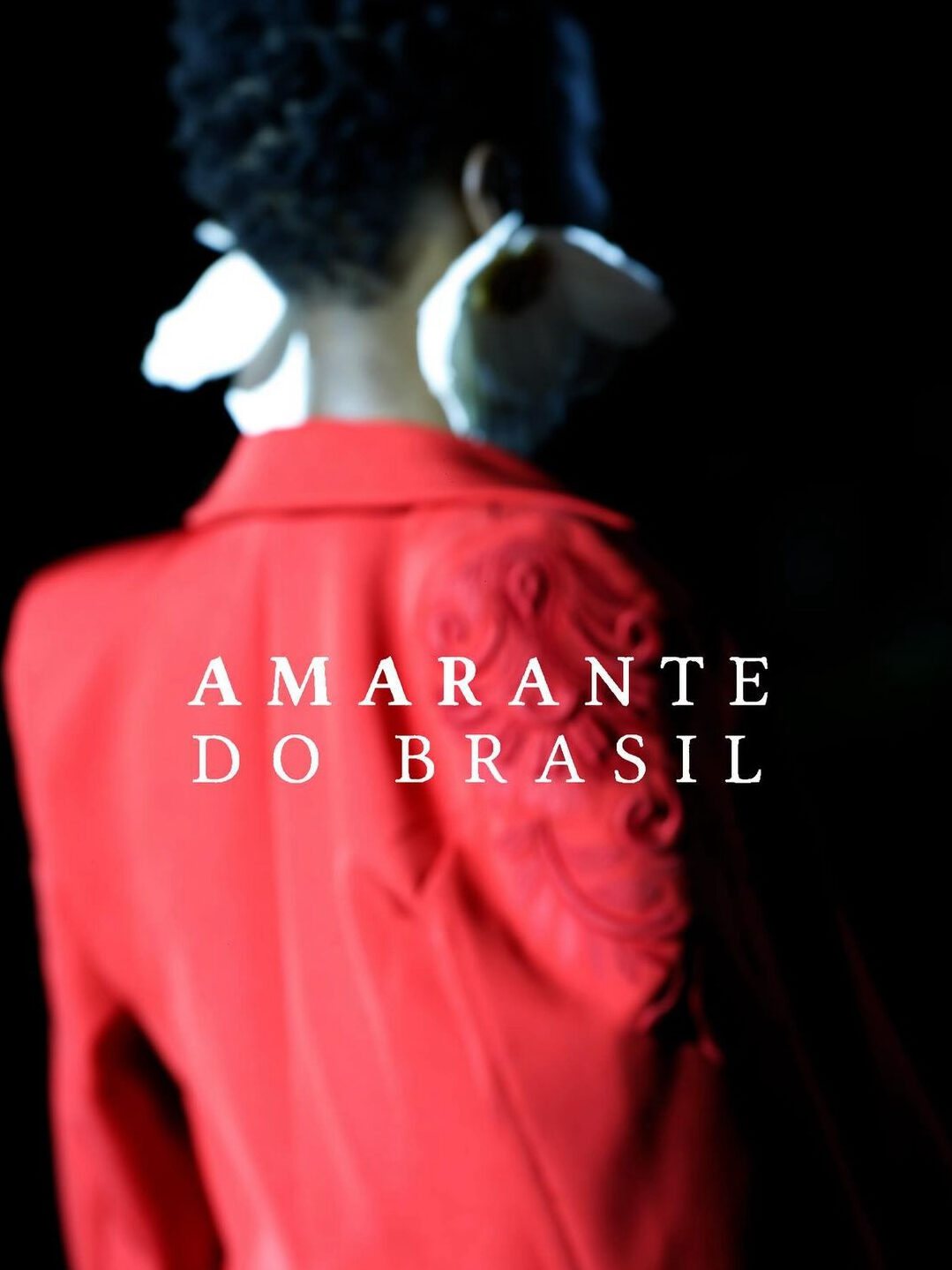 Fotografia de moda de modelo vestindo um casaco vermelho e brincos de flor grandes em cor branca. Por cima da foto, há a aplicação da frase "Amarante do Brasil".