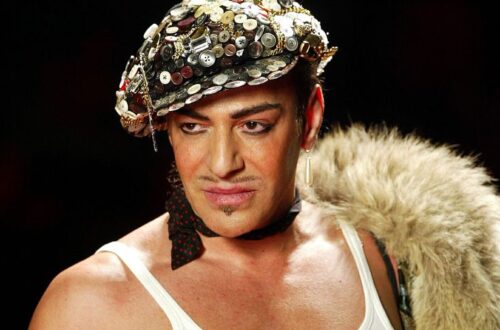 Foto do designer britânico Jonh Galliano usando uma boina de botões e outras texturas, uma regata branca sendo fotografato em plano médio. O designer britânico John Galliano aparece na passarela ao final de sua coleção de alta-costura Primavera/Verão 2003 para a maison Dior, em Paris, em 20 de janeiro de 2003.