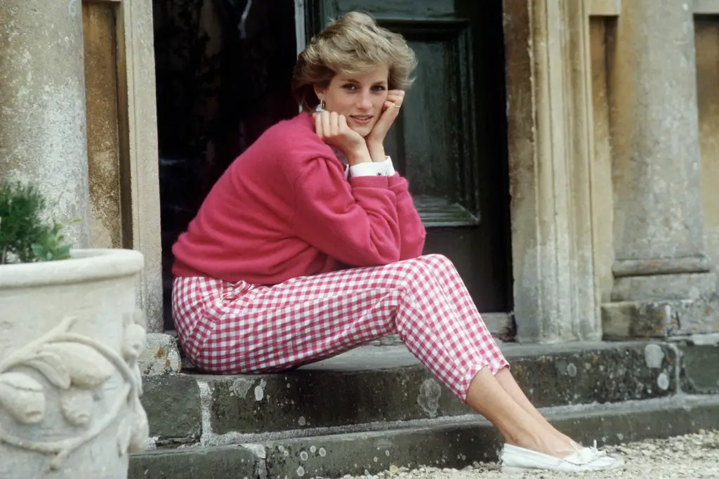 Diana aparece sentada em uma escada em frente a uma porta que dá para a rua vestindo um suéter e uma calça Capri xadrez, ambos em rosa, transmitindo proximidade e fugindo do ideal de rigidez e glamour que normalmente associamos aos membros da realeza.